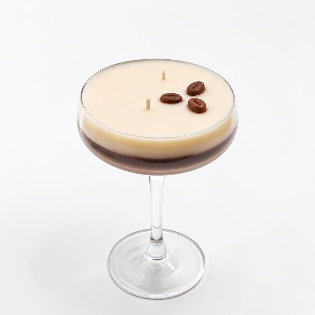 A layered candle in a coupe glass, featuring a creamy beige top layer and a dark brown bottom layer, with three coffee beans and two wicks on top, set against a plain white background.