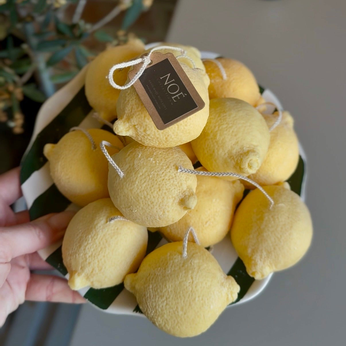 A hand holds a white bowl filled with yellow lemon-shaped bath bombs or bath toppers, with a small hanging tag reading 'NOE'. The background shows a gray tabletop, a brick wall, and a candle.