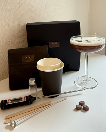 A candle-making kit and finished product displayed on a white table against a beige wall. The kit includes black boxes with a gold NOE logo, a black vessel, a double-wick candle in a coupe glass, a digital thermometer, wooden sticks, wicks, and small chocolate pieces.