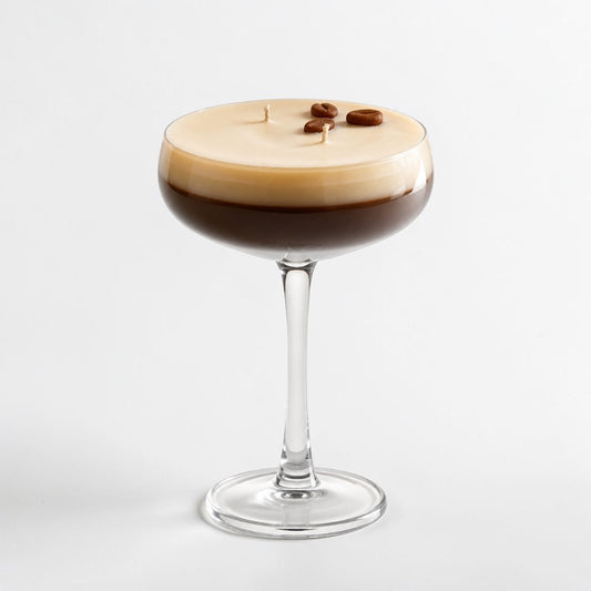A stemmed coupe glass candle filled with a layered coffee-inspired design, featuring a dark brown lower section and a thick beige cream-colored top layer with three small coffee beans on top, set against a plain light gray background.