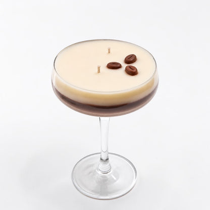 A layered candle in a coupe glass, featuring a creamy beige top layer and a dark brown bottom layer, with three coffee beans and two wicks on top, set against a plain white background.
