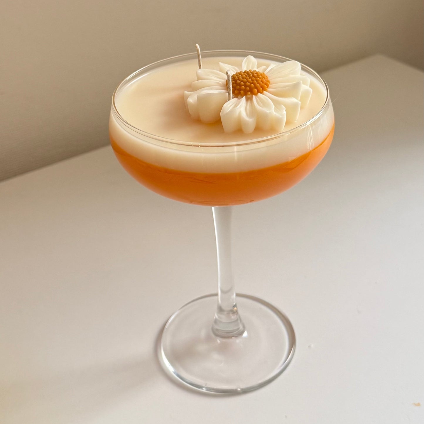 A layered cocktail candle in a coupe glass, featuring a frothy white top layer with a piped daisy flower and small candle wick, over an orange-brown bottom layer, set on a white surface against a beige wall.