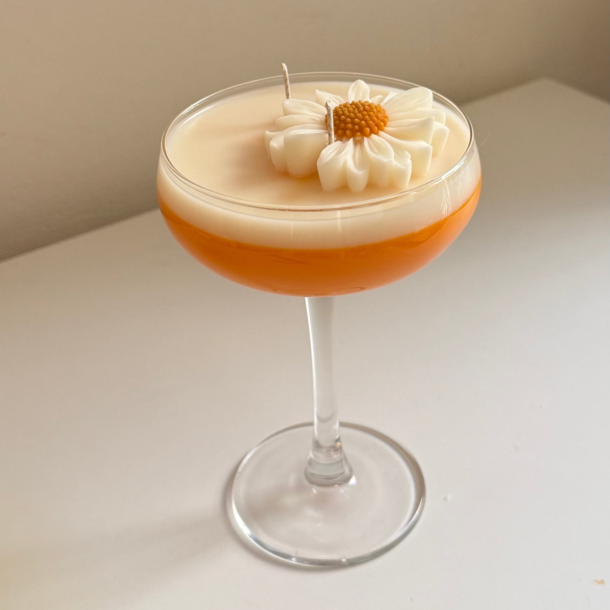 A layered cocktail candle in a coupe glass, featuring a frothy white top layer with a piped daisy flower and small candle wick, over an orange-brown bottom layer, set on a white surface against a beige wall.
