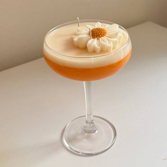 A layered cocktail candle in a coupe glass, featuring a frothy white top layer with a piped daisy flower and small candle wick, over an orange-brown bottom layer, set on a white surface against a beige wall.