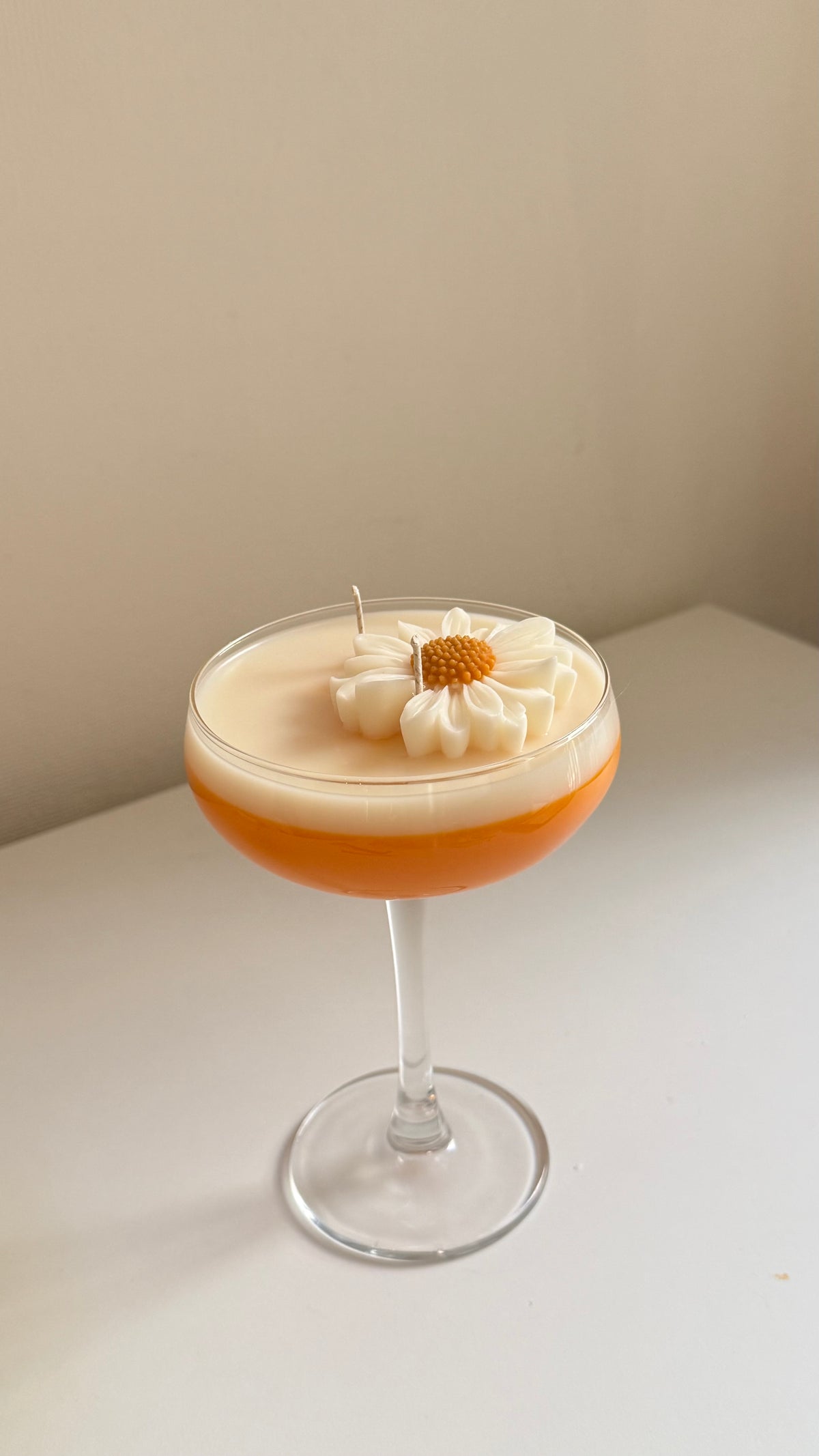 A stemmed glass candle with a layered design, featuring a warm orange base and a thick cream-colored top layer decorated with a white daisy flower and a small wick, set on a white surface against a beige wall.