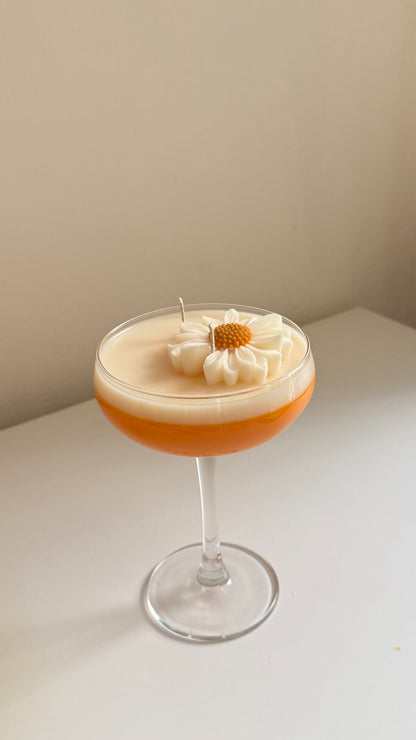 A stemmed glass candle with a layered design, featuring a warm orange base and a thick cream-colored top layer decorated with a white daisy flower and a small wick, set on a white surface against a beige wall.