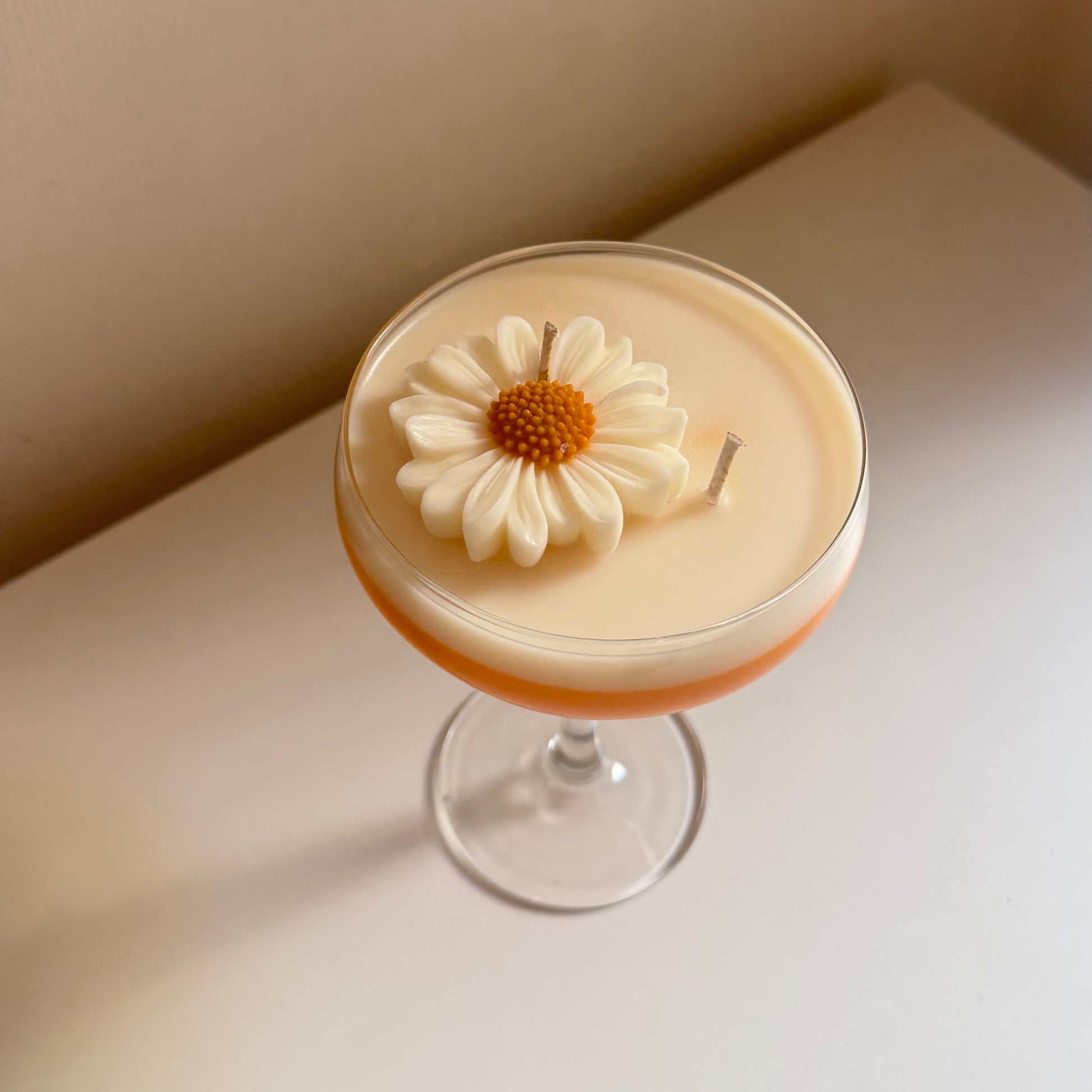 A low-angle shot of a cocktail in a coupe glass, featuring a creamy beige body with a thin orange layer at the bottom and a white daisy flower garnish on top, set on a white surface against a beige wall.