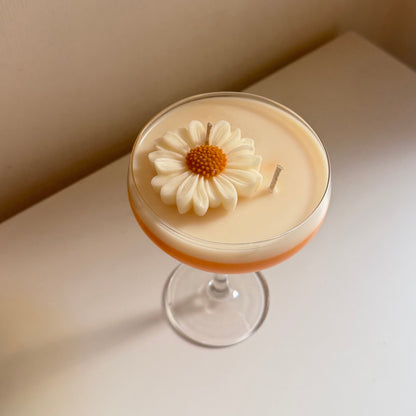A low-angle shot of a cocktail in a coupe glass, featuring a creamy beige body with a thin orange layer at the bottom and a white daisy flower garnish on top, set on a white surface against a beige wall.